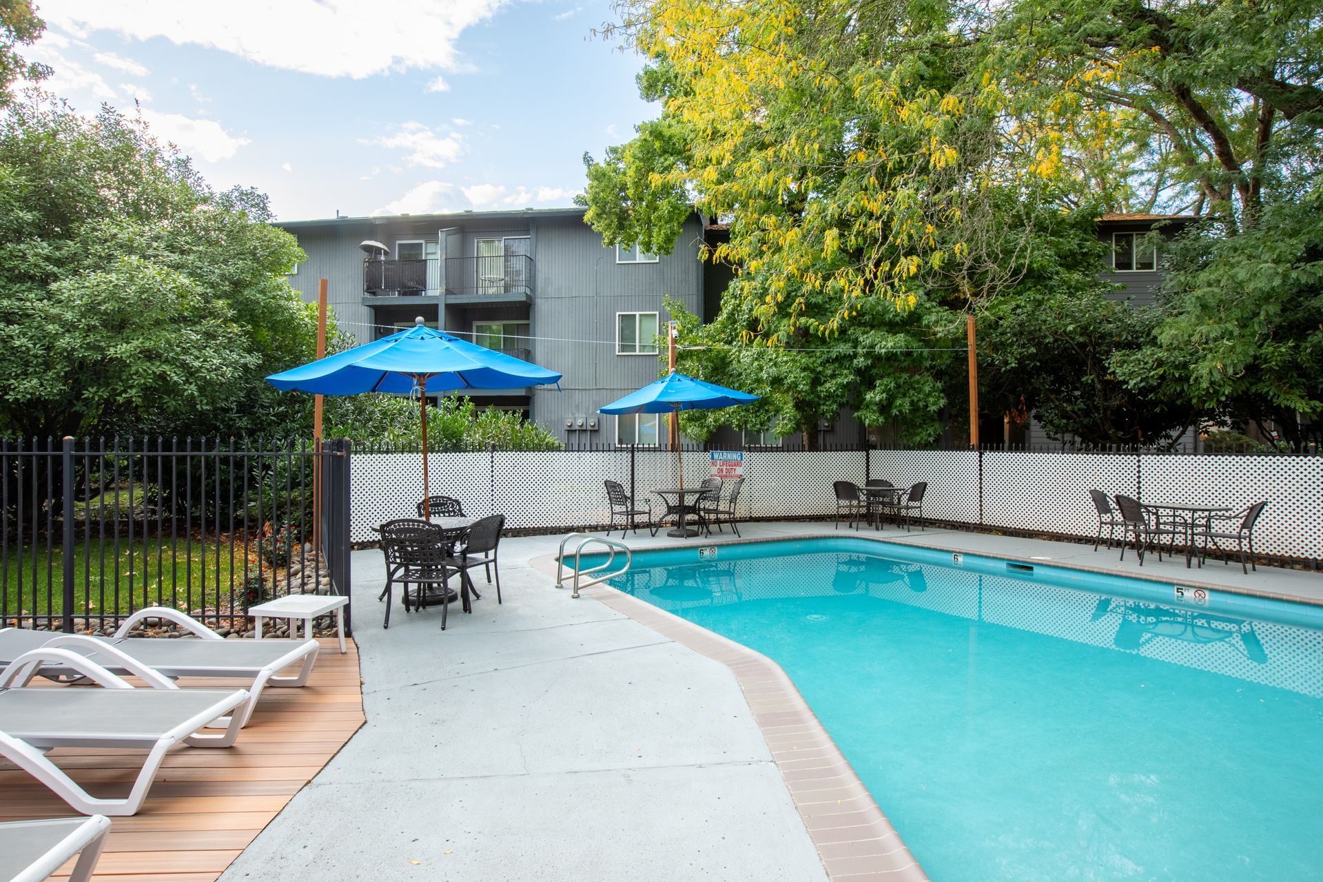 Pool area with blue umbrellas, tables, and chairs next to an apartment building.