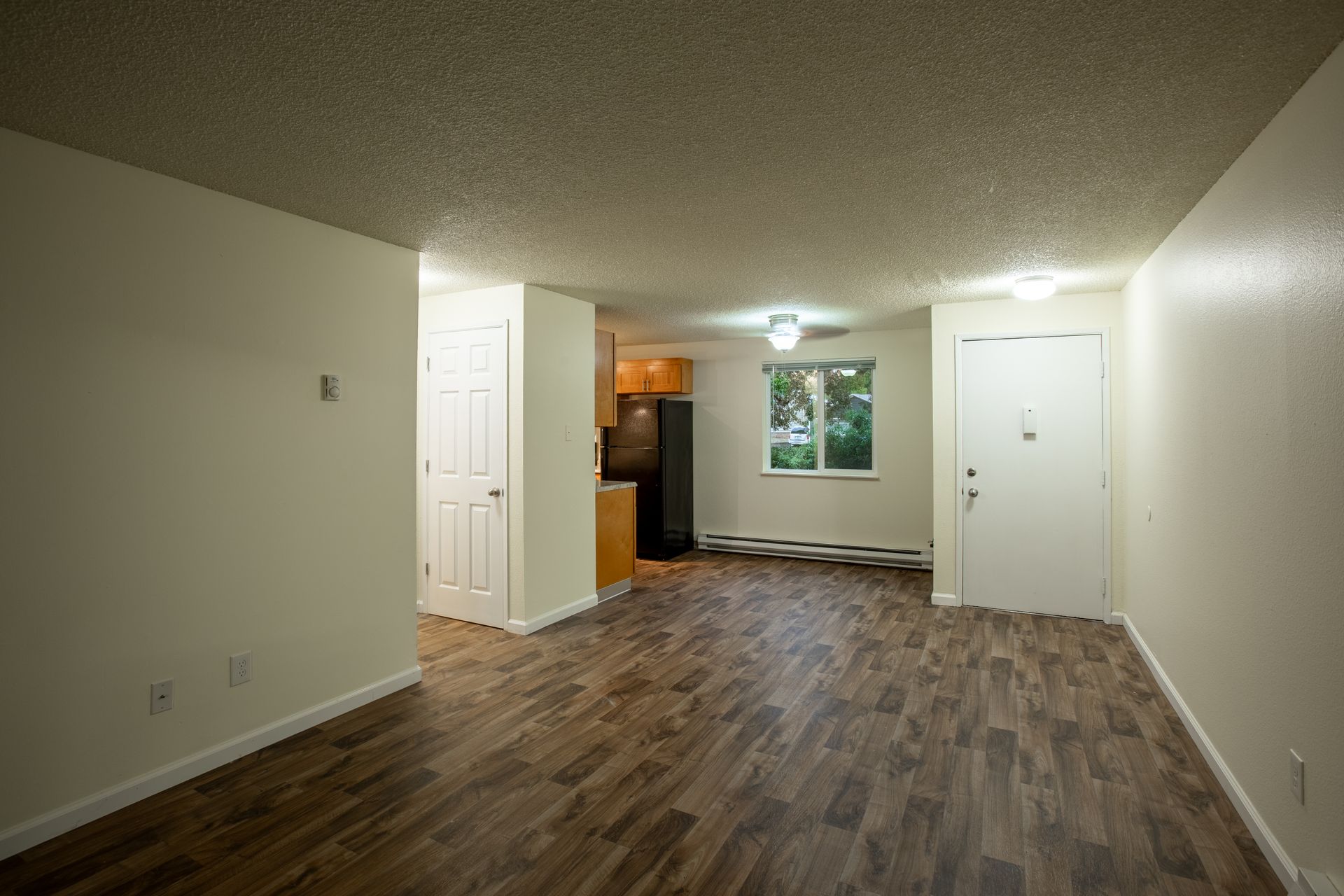 Interior view of an apartment with wooden floors, white walls, a kitchen in the back, and a window.