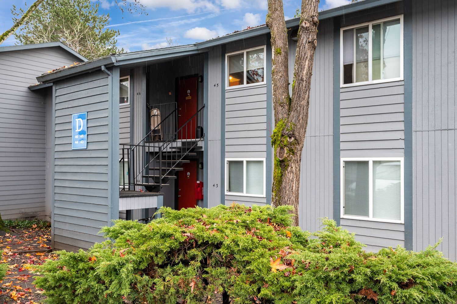 Gray apartment building with a red door, surrounded by green bushes and a tree.