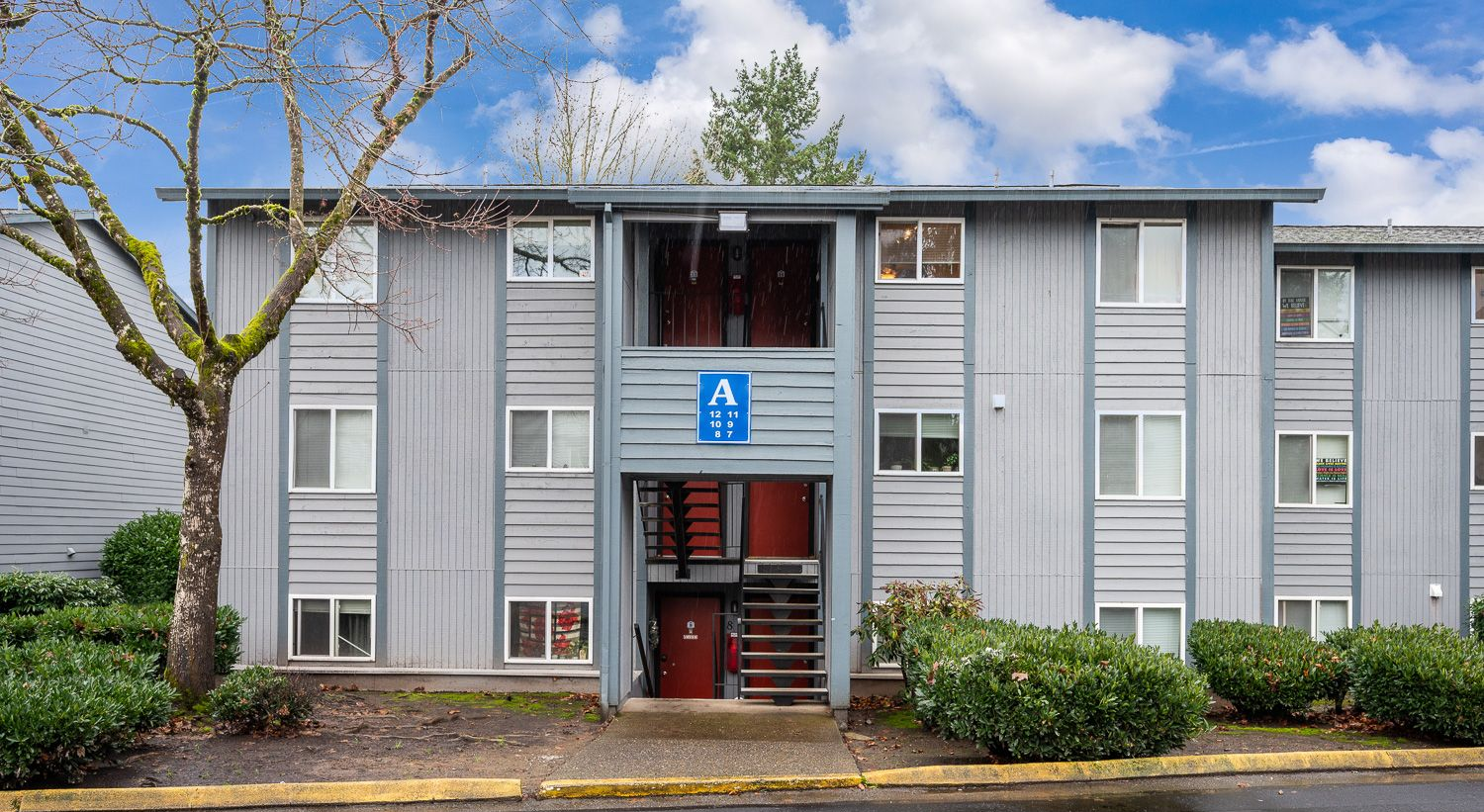 Apartment building exterior with gray siding, a central stairwell, and green shrubs.