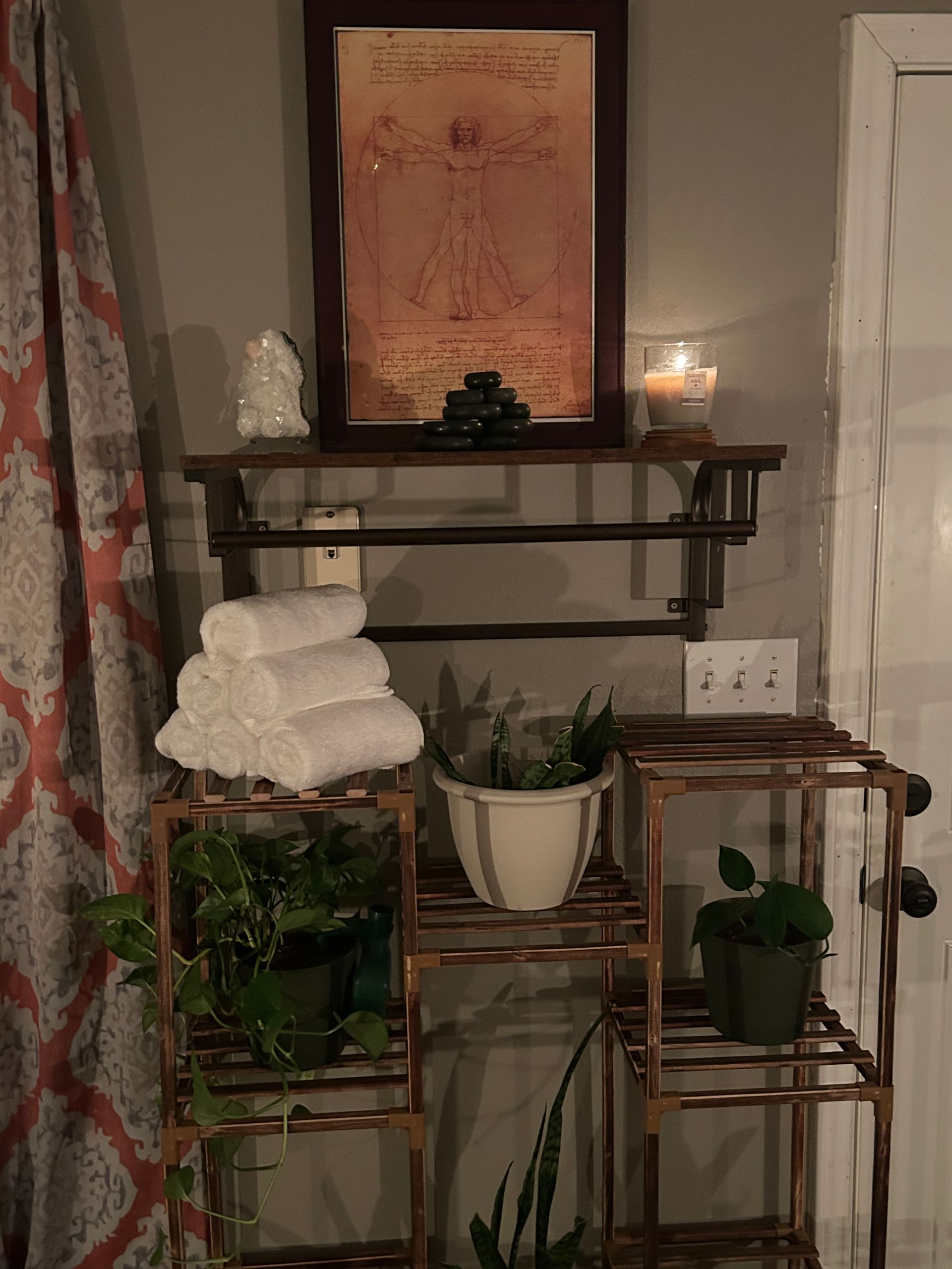 A wooden shelf with a framed drawing, candle, stacked towels, and potted plants.