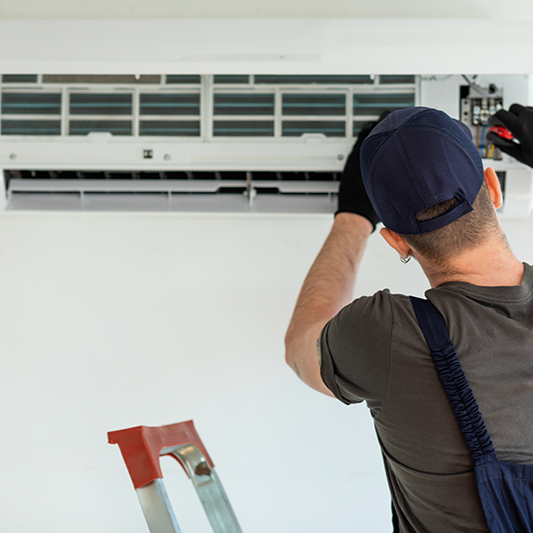 Un technicien en casquette et uniforme répare un climatiseur mural blanc, debout sur une échelle.