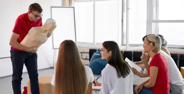 A man is teaching a group of people how to do a heart massage on a mannequin.