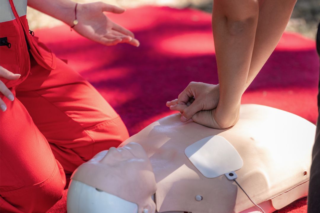 A person practices CPR on a medical training mannequin while an instructor in red clothing looks on.