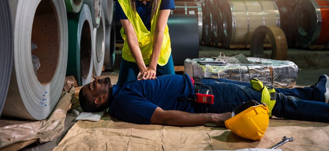 Person performing CPR on a man in a warehouse. The man is unconscious. Metal rolls are in the background.