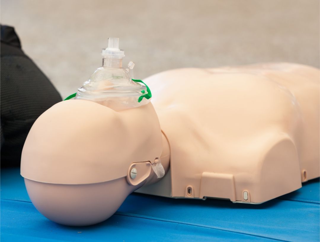 A CPR training mannequin lying on a blue surface with an oxygen mask placed over its nose and mouth.