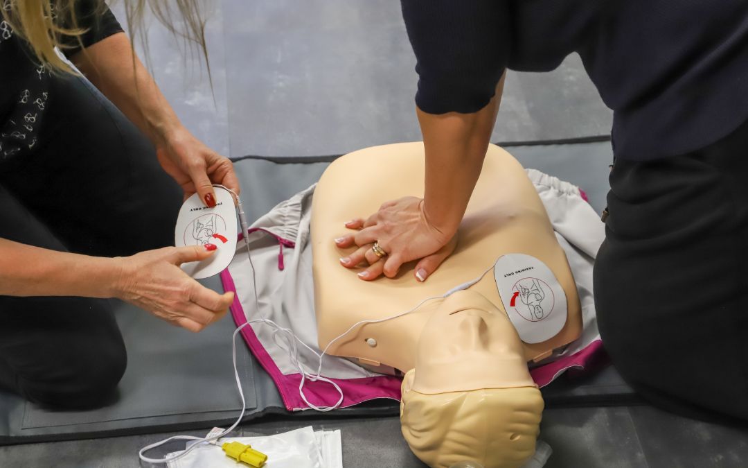 Two people practice CPR on a medical mannequin, one performing chest compressions and the other preparing AED pads.