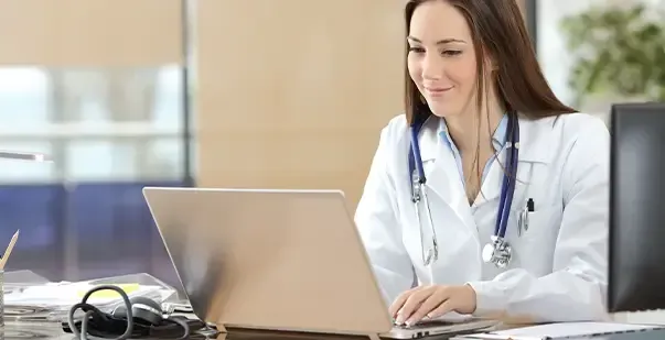 A female doctor is sitting at a desk using a laptop computer.