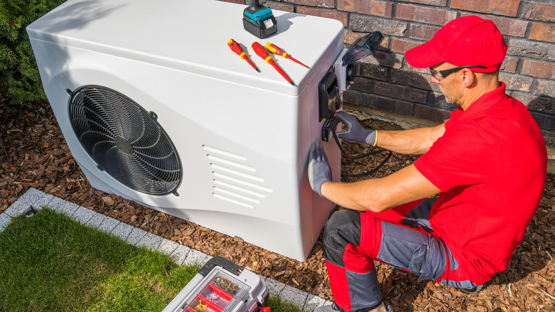 Technician in red uniform repairing an outdoor HVAC unit beside a house with tools nearby