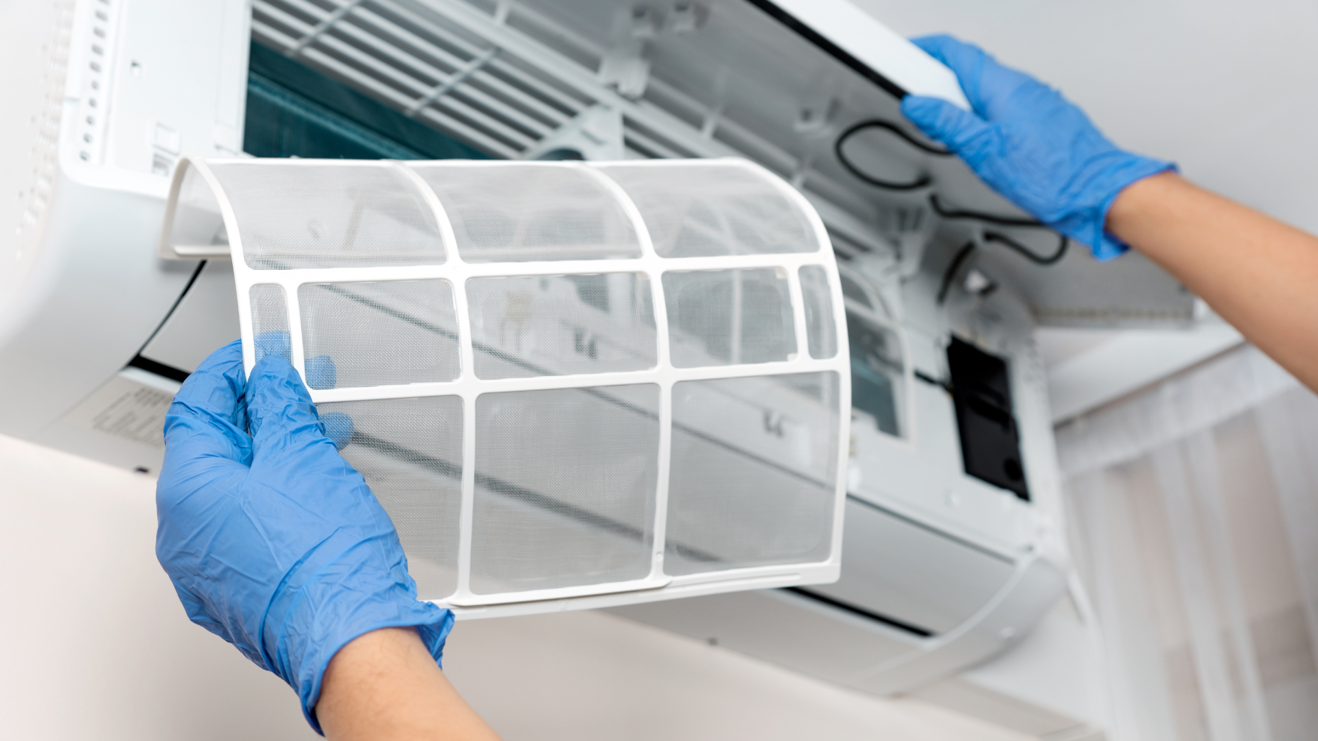 Blue-gloved hands removing a white air conditioner filter from a wall-mounted unit
