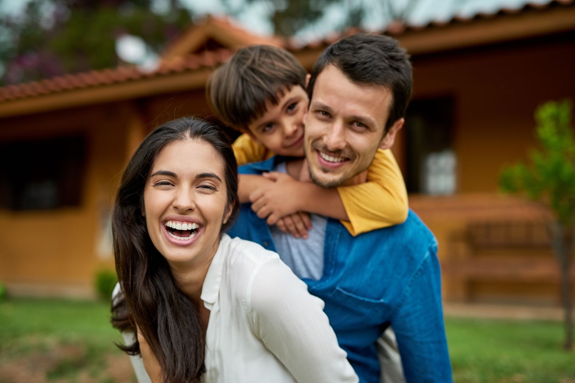 A man and woman are giving a child a piggyback ride in front of a house.