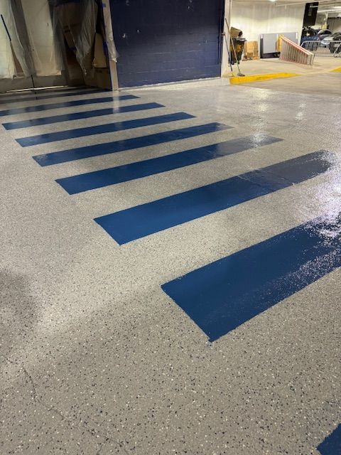 A pedestrian crosswalk with blue painted stripes on a light-colored, speckled epoxy garage floor.