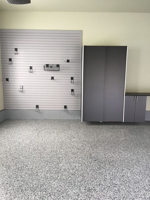 A garage interior with gray speckled flooring, a slat wall organizer with hooks, and dark gray cabinets with a countertop.