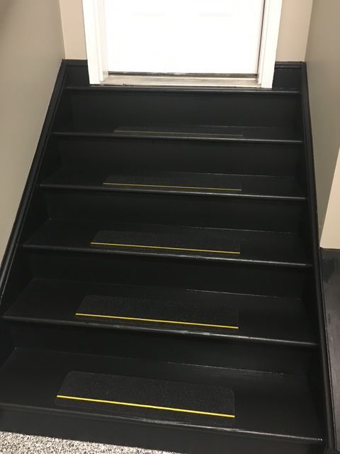 A straight wooden staircase painted black with yellow anti-slip adhesive strips on each step leading up to a closed door.