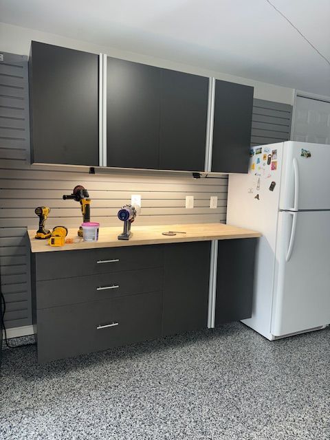 A garage workspace with dark gray cabinets, a light wood countertop, wall-mounted slat boards, and a white refrigerator.