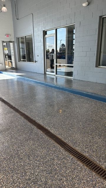 A view of a building exterior with grey block walls, large glass windows and doors, and a speckled floor with a drain.