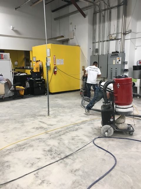 A worker uses an industrial floor sander and vacuum in a large room with a prominent yellow wall.