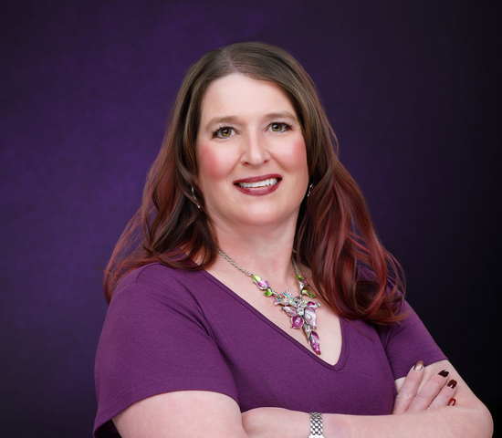 Woman with auburn hair in purple top smiling with arms crossed; purple background.