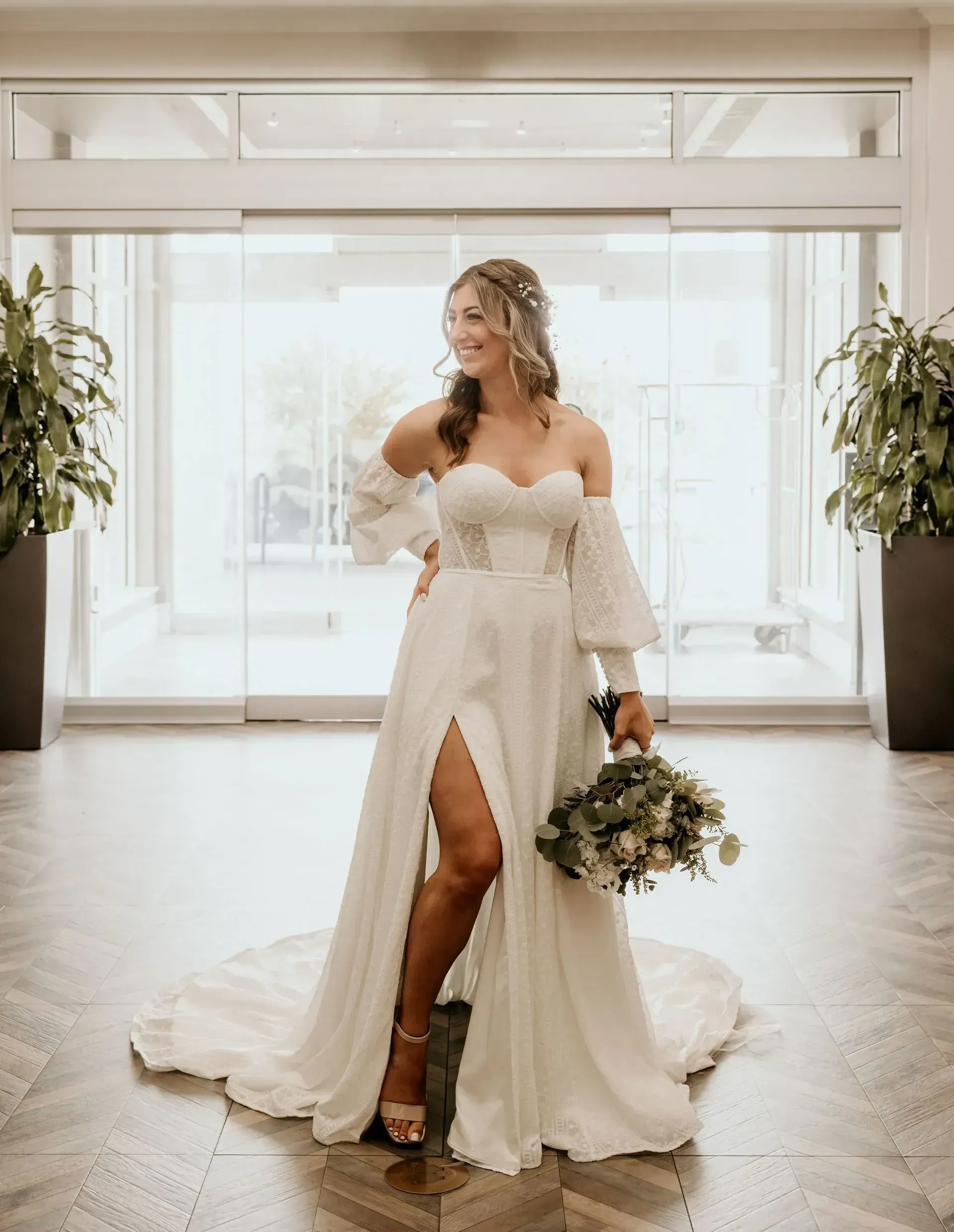 A bride in a white dress is standing in a hallway holding a bouquet of flowers.