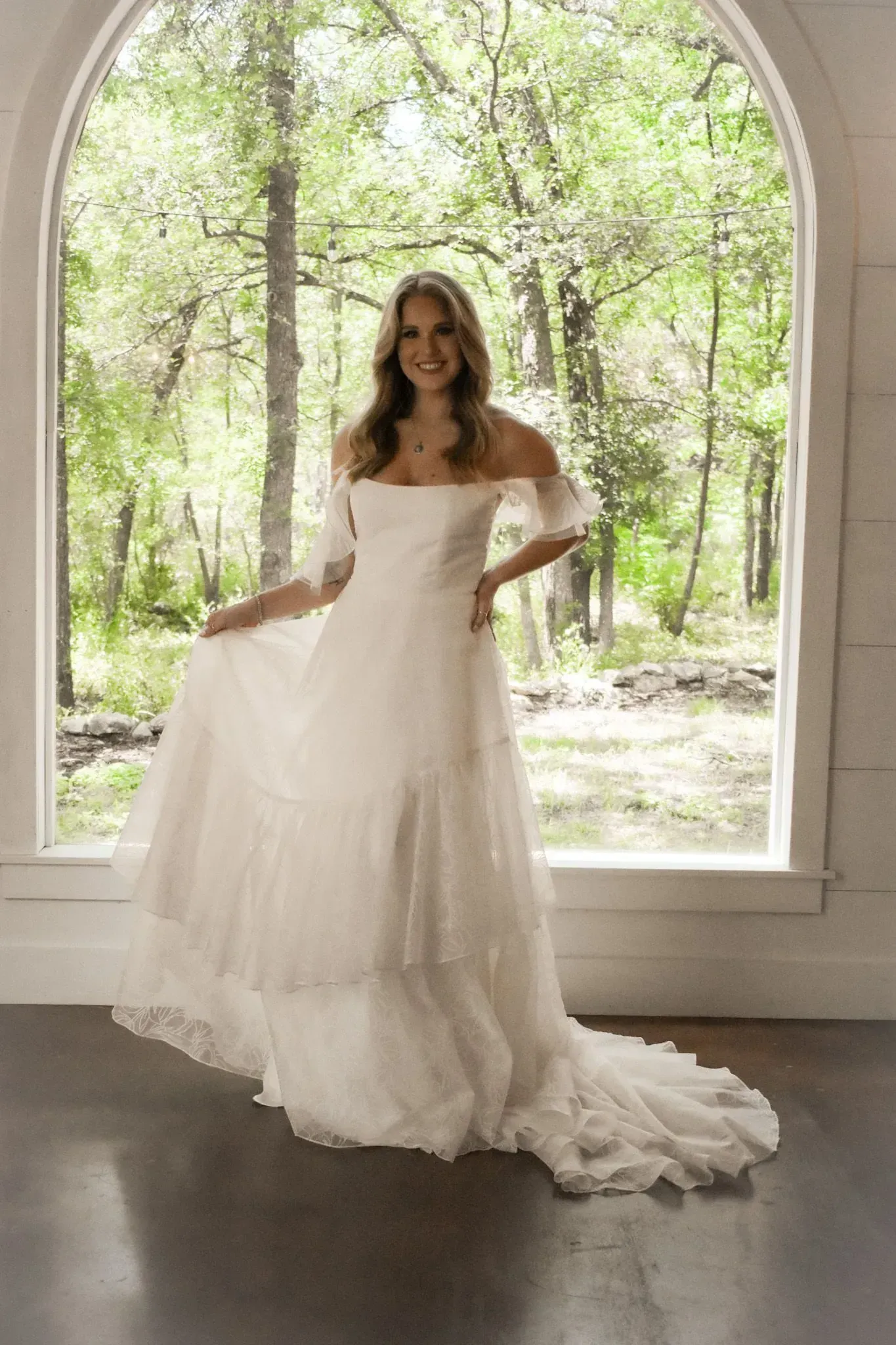 A woman in a white wedding dress is standing in front of a window.