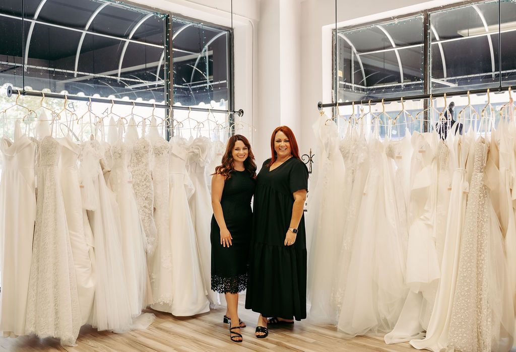 Two women are standing next to each other in front of a wall of wedding dresses.