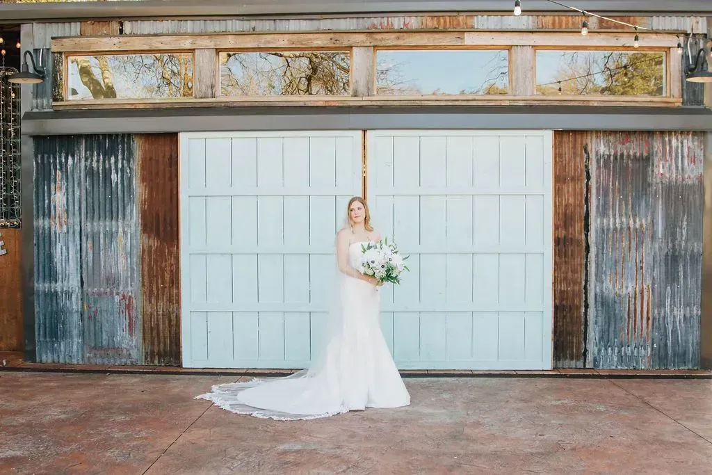 A bride in a wedding dress is standing in front of a garage door holding a bouquet of flowers.