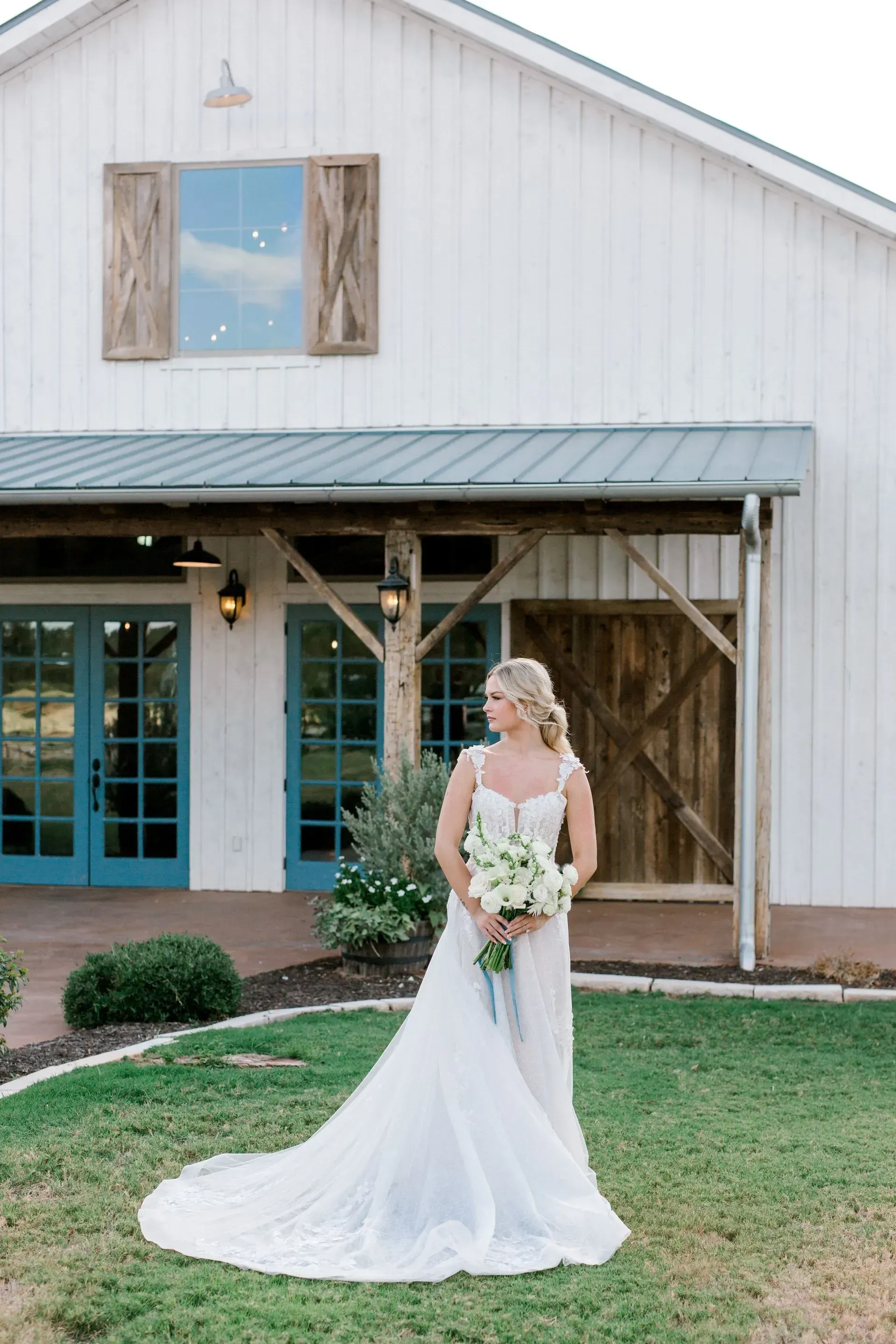 A bride in a wedding dress is standing in front of a white barn holding a bouquet of flowers.