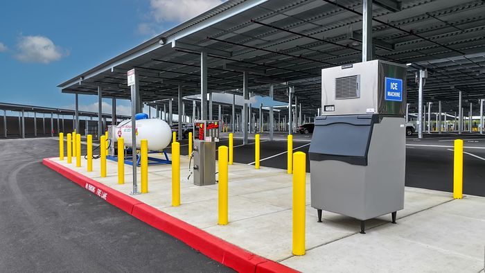 Covered outdoor industrial area with yellow bollards, a propane tank, and a gray machine on a concrete pad.