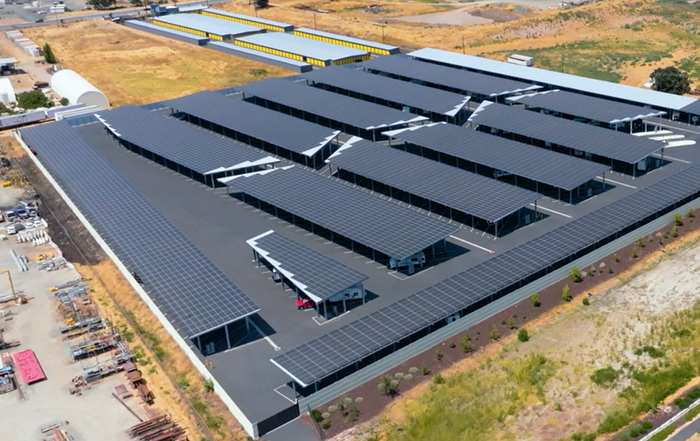 Aerial view of a large industrial building with rows of rooftop solar panels and surrounding dry land