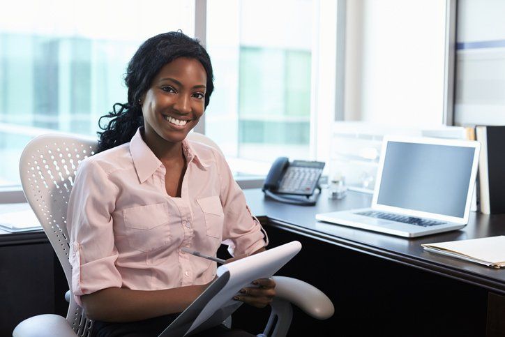A woman smiling in front of her desk holding a notepad