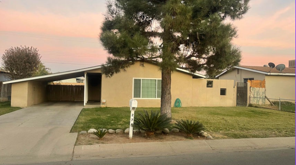A single-story beige house with a covered carport, a tree in front, and a green lawn under a pink sky.
