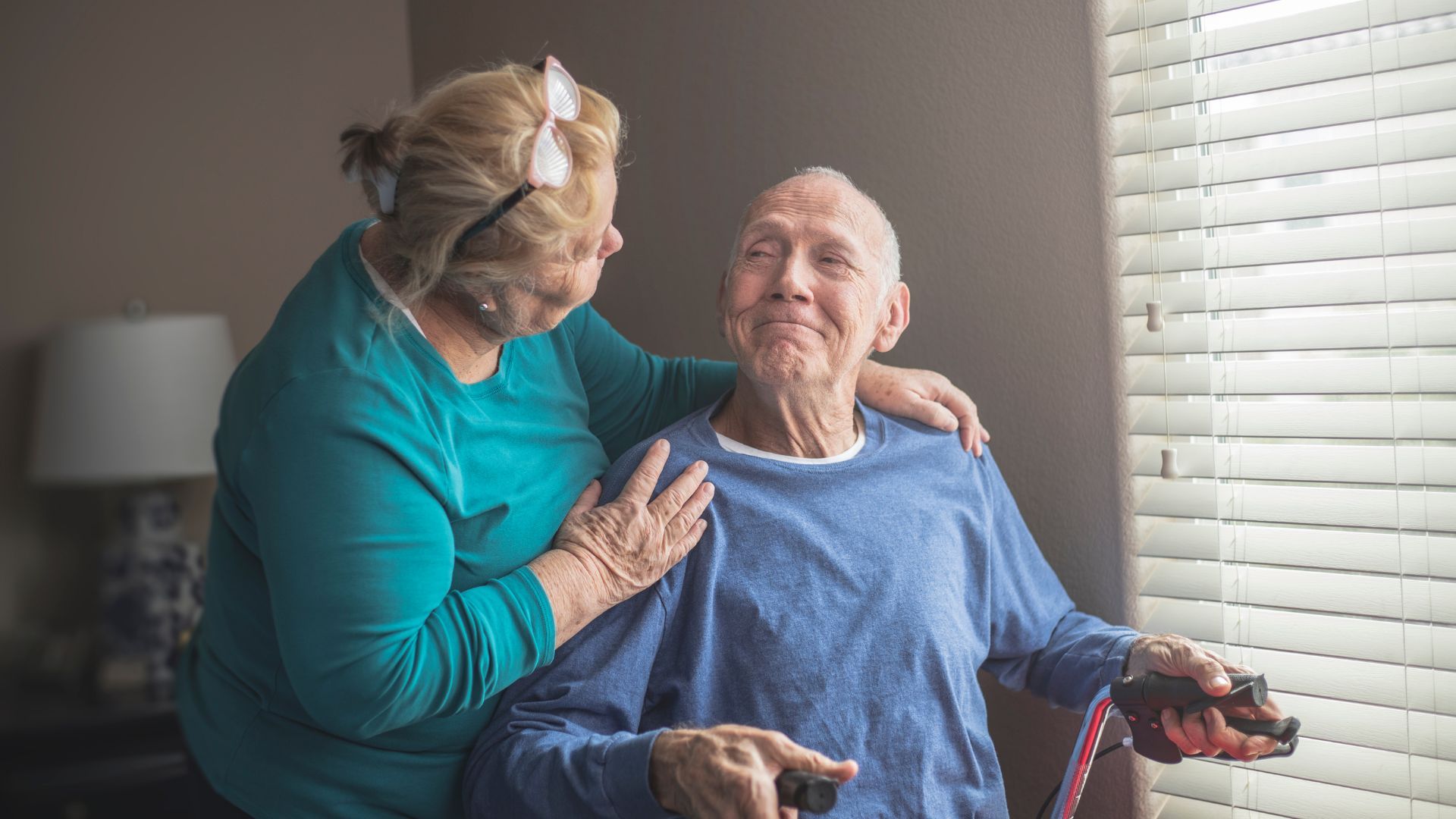 A person in a blue shirt holds a remote while being supported by someone in a teal sweater indoors near a window.