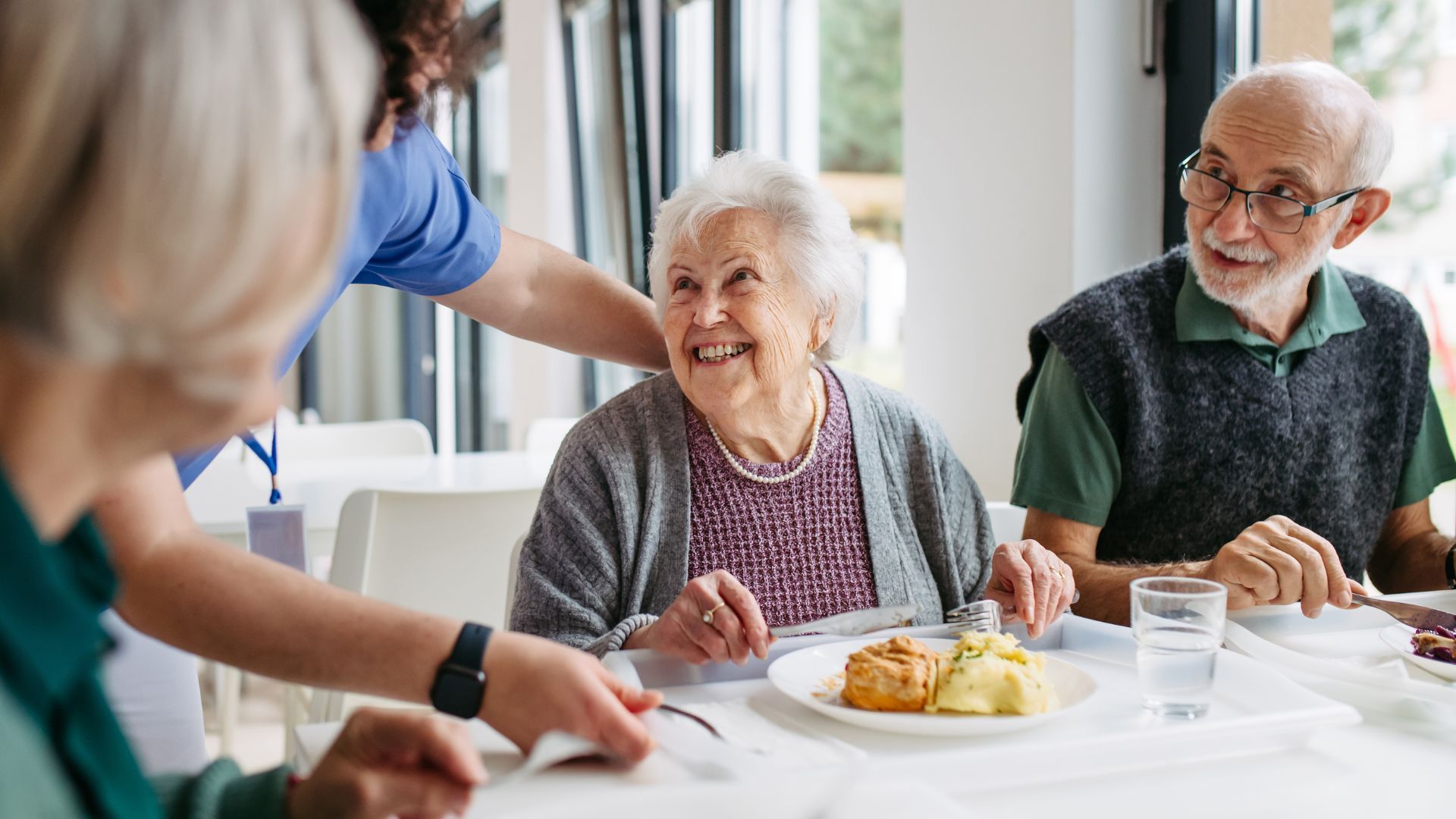 A caregiver assisting an elderly person with a meal in a bright dining room, while another person watches nearby.