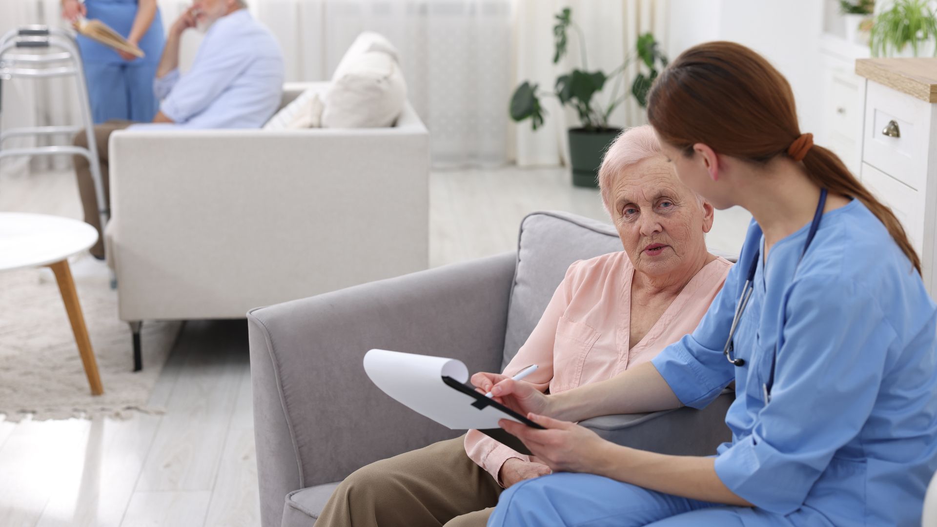 A healthcare worker in blue scrubs sits on a sofa, reviewing notes with an older patient in a bright, modern living space.