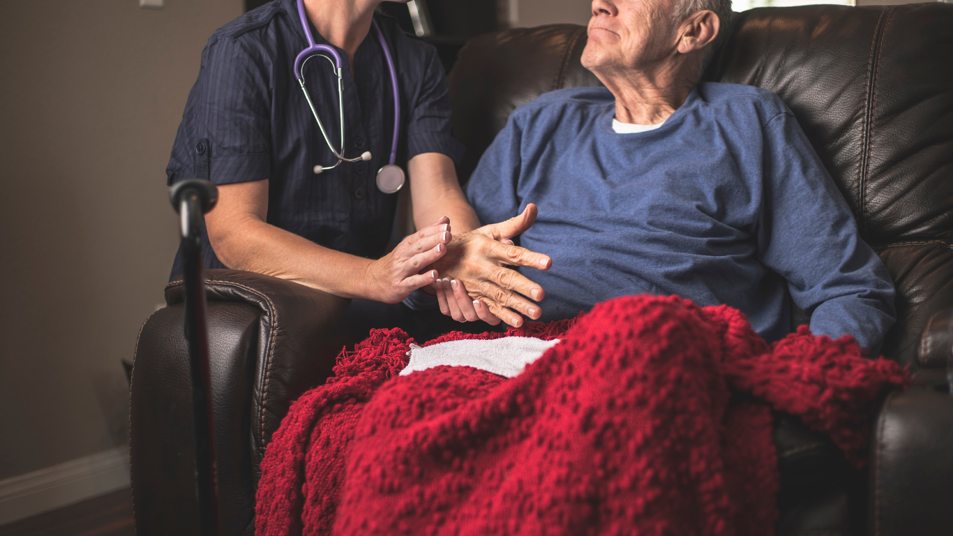 A person in medical scrubs holding an older person's hand as they sit together on a leather sofa covered by a red blanket.