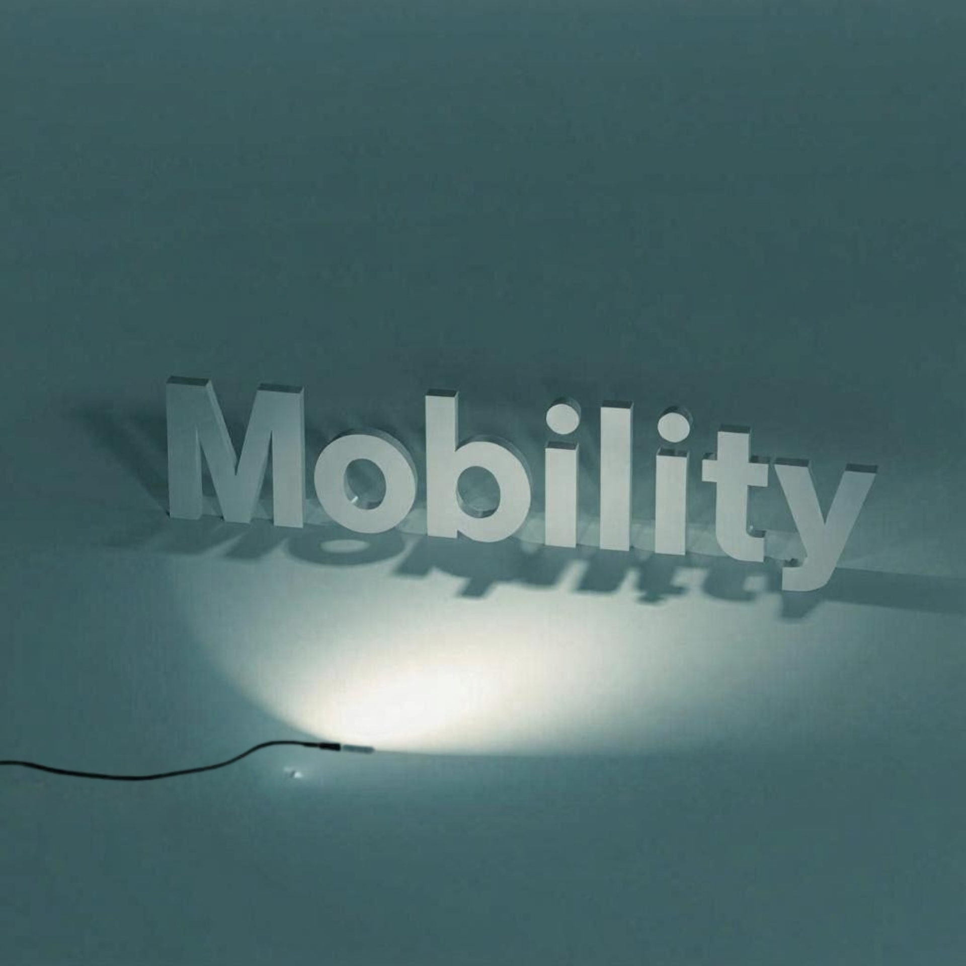 „Mobilität“ in 3D-Buchstaben, von unten beleuchtet, wirft einen Schatten auf eine gedämpfte blaugrüne Oberfläche.
