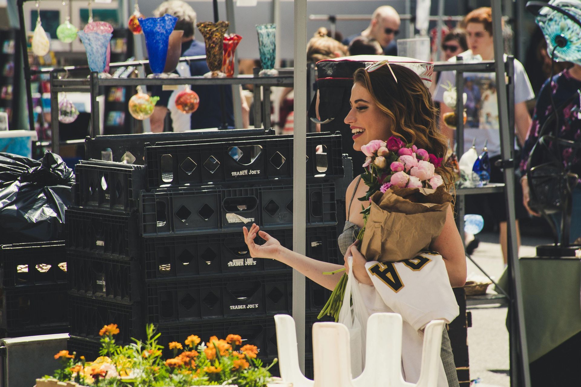 Frau mit Blumen in der Hand lächelt auf einem Markt und betrachtet einen Gegenstand. Viele Menschen haben Waren im Blick.