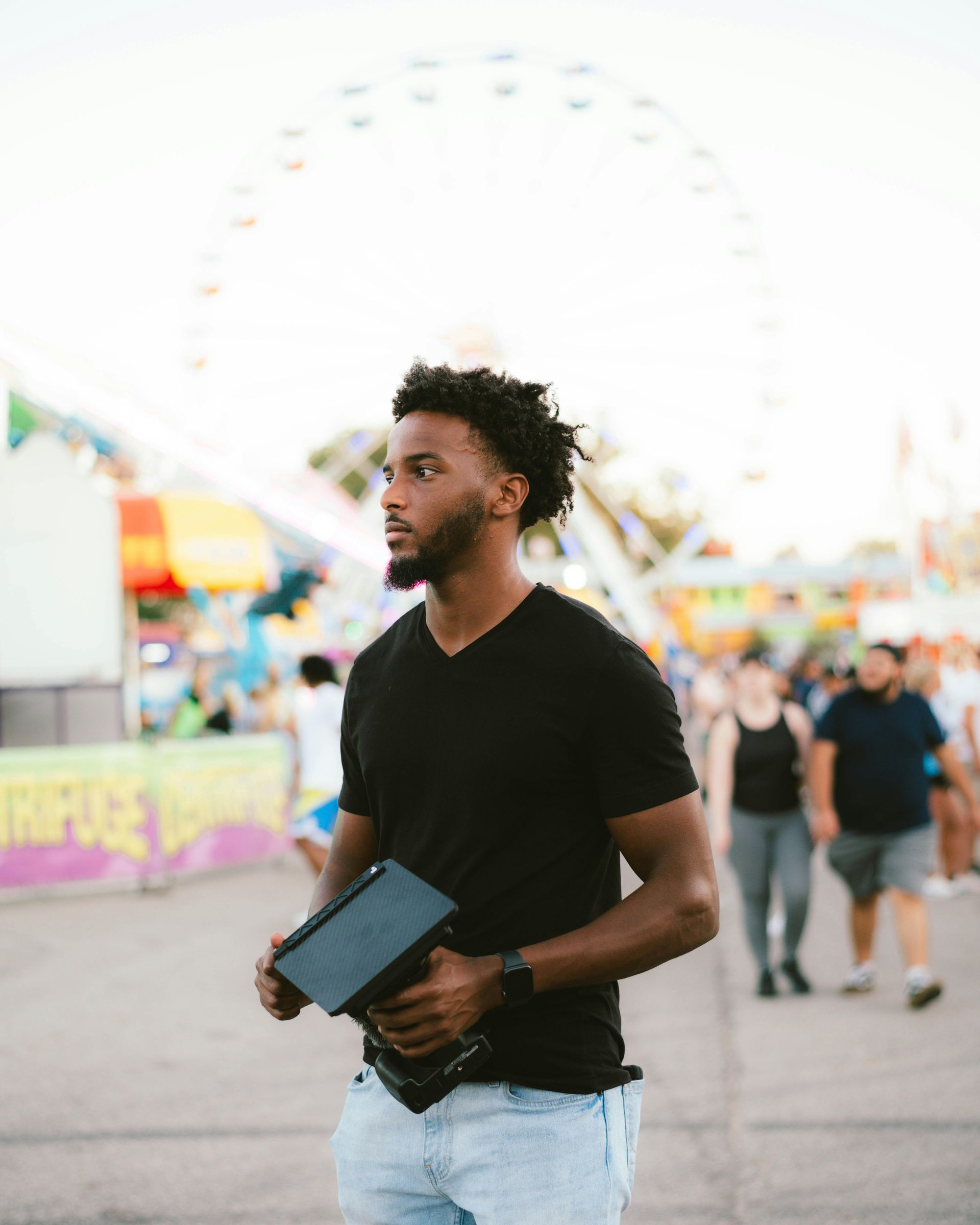 Ein Mann in schwarzem Hemd hält auf einem Jahrmarkt ein Tablet in der Hand. Im Hintergrund ist ein Riesenrad zu sehen.