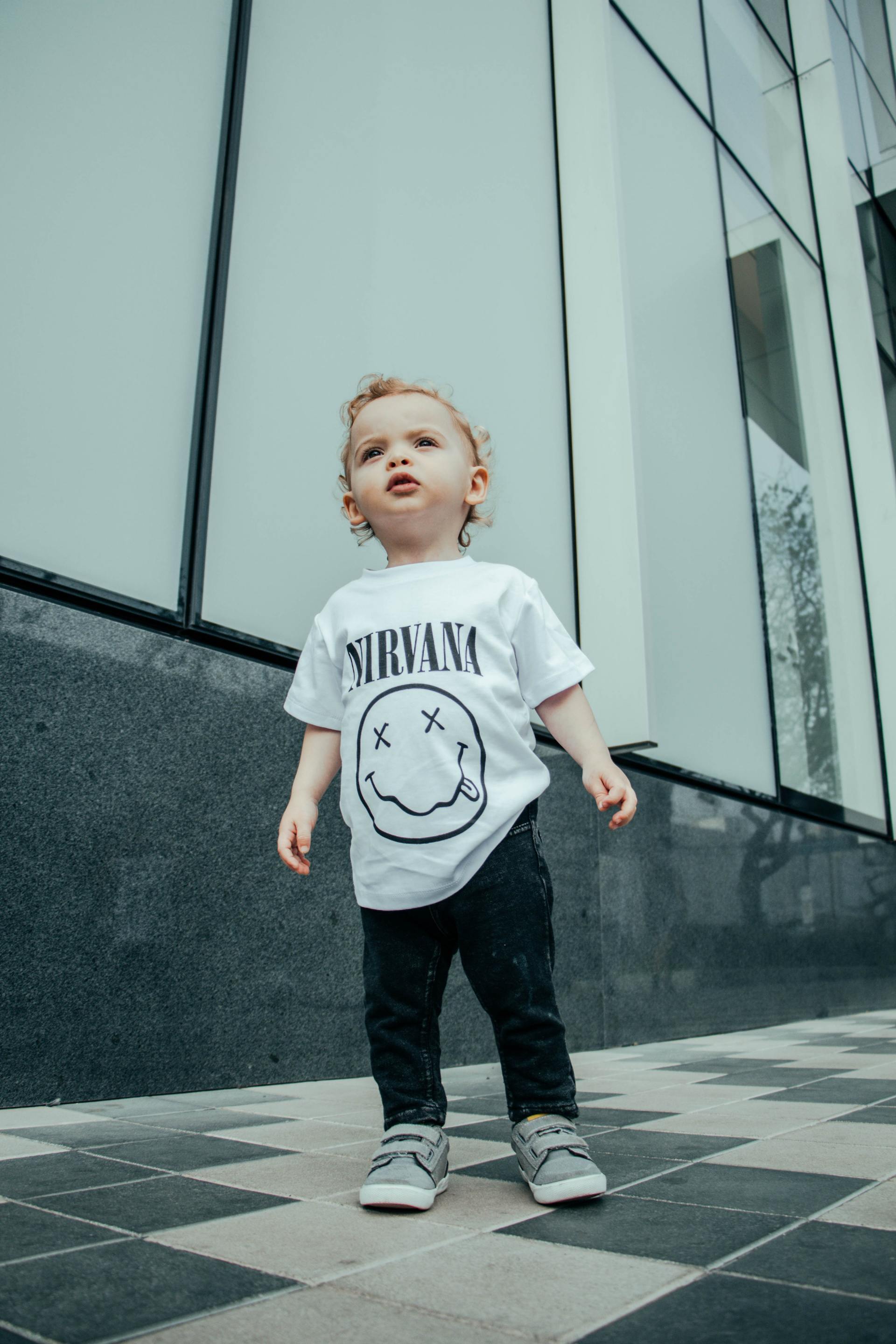 A young child wearing a Nirvana t-shirt and black pants stands outdoors, looking up.
