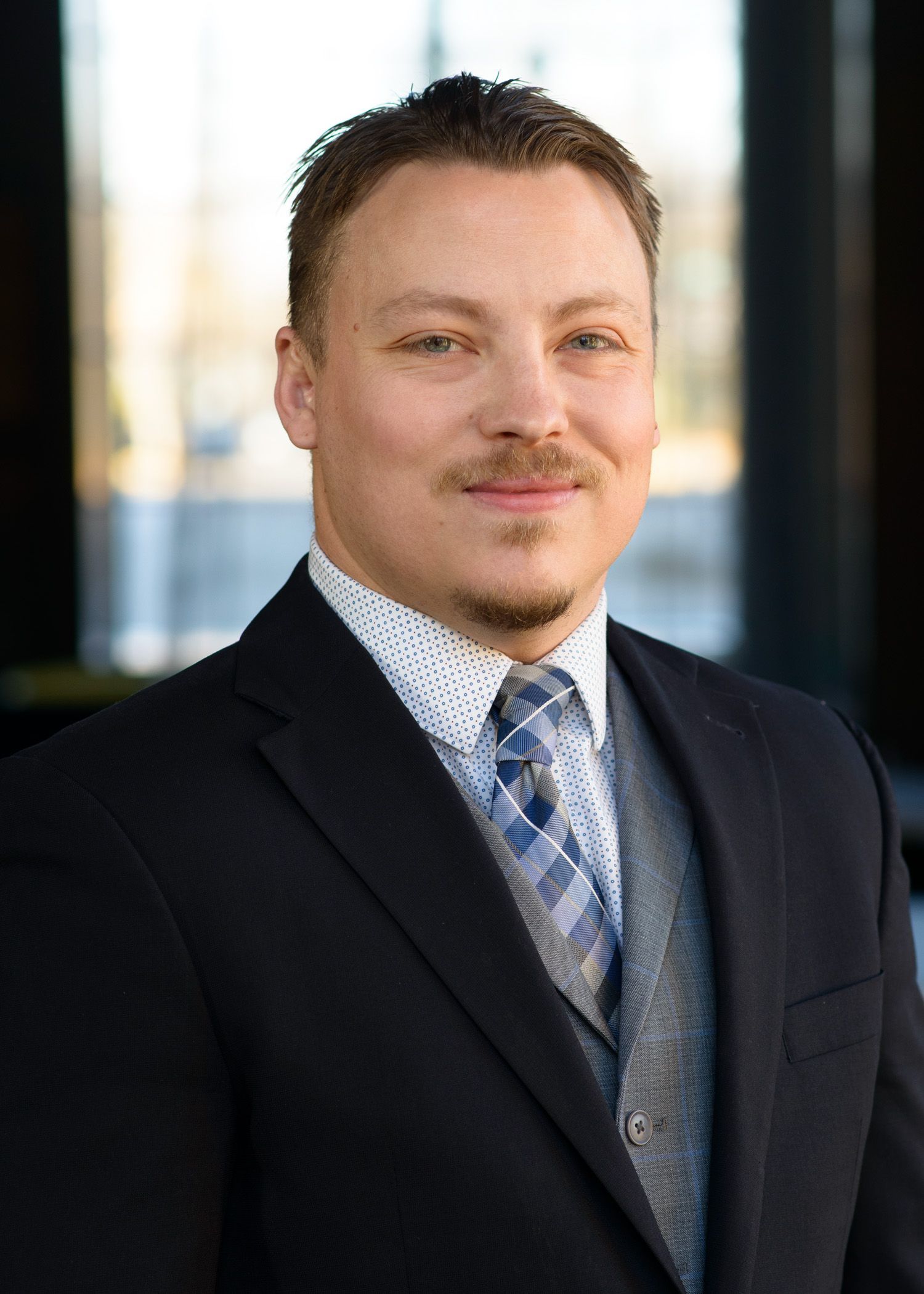 Man in suit smiles, indoors; patterned tie, vest, blurred background.