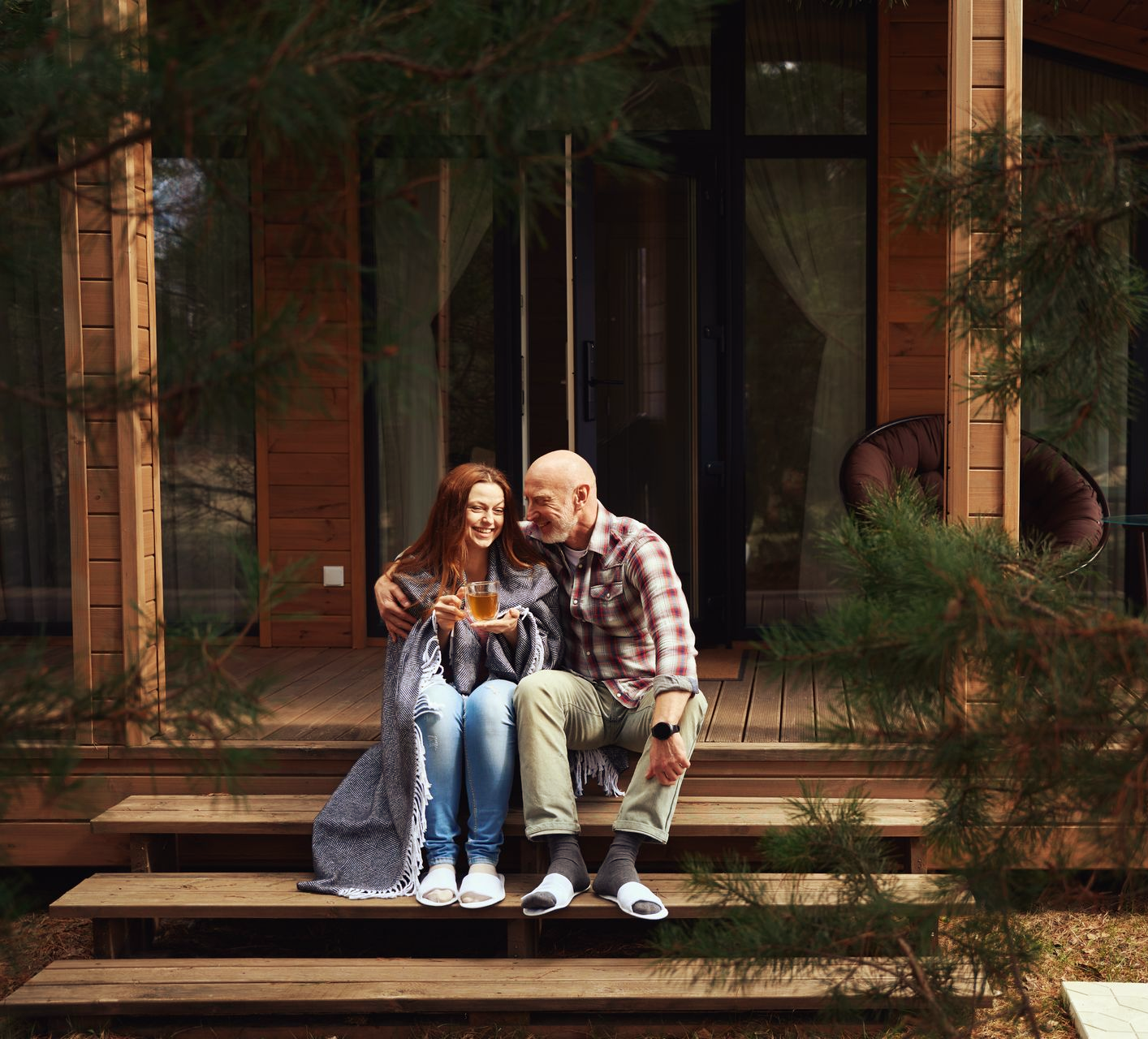 Couple wrapped in blanket, smiling, sitting on wooden steps in front of cabin, holding drinks.