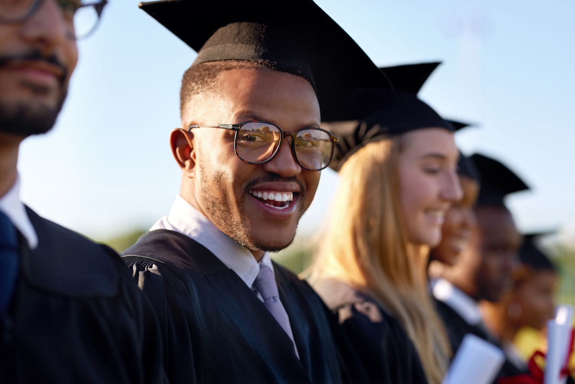 Graduation: Smiling graduate in cap and gown, looking at camera, other grads in background.