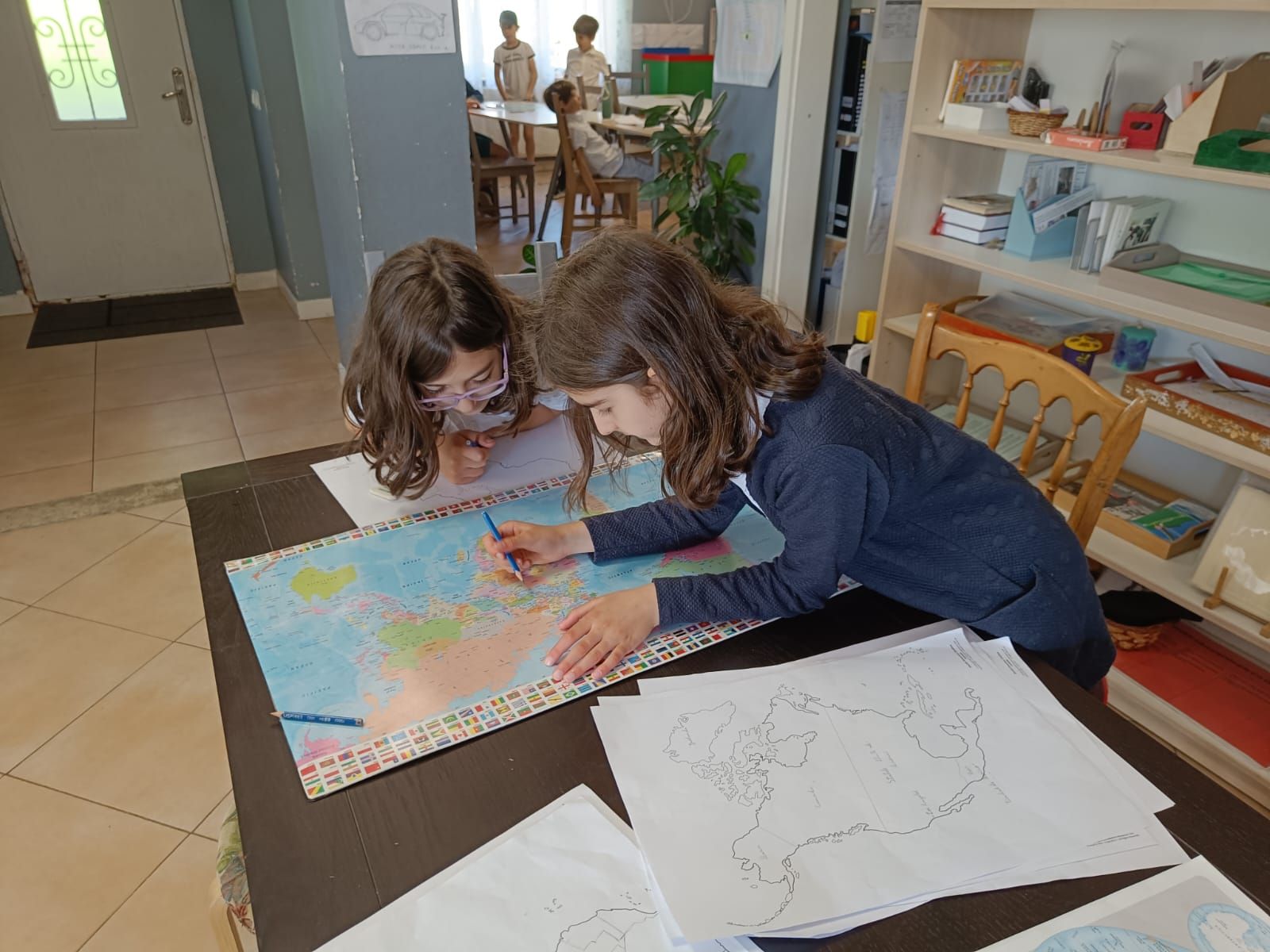 Two young girls are sitting at a table drawing on a piece of paper.