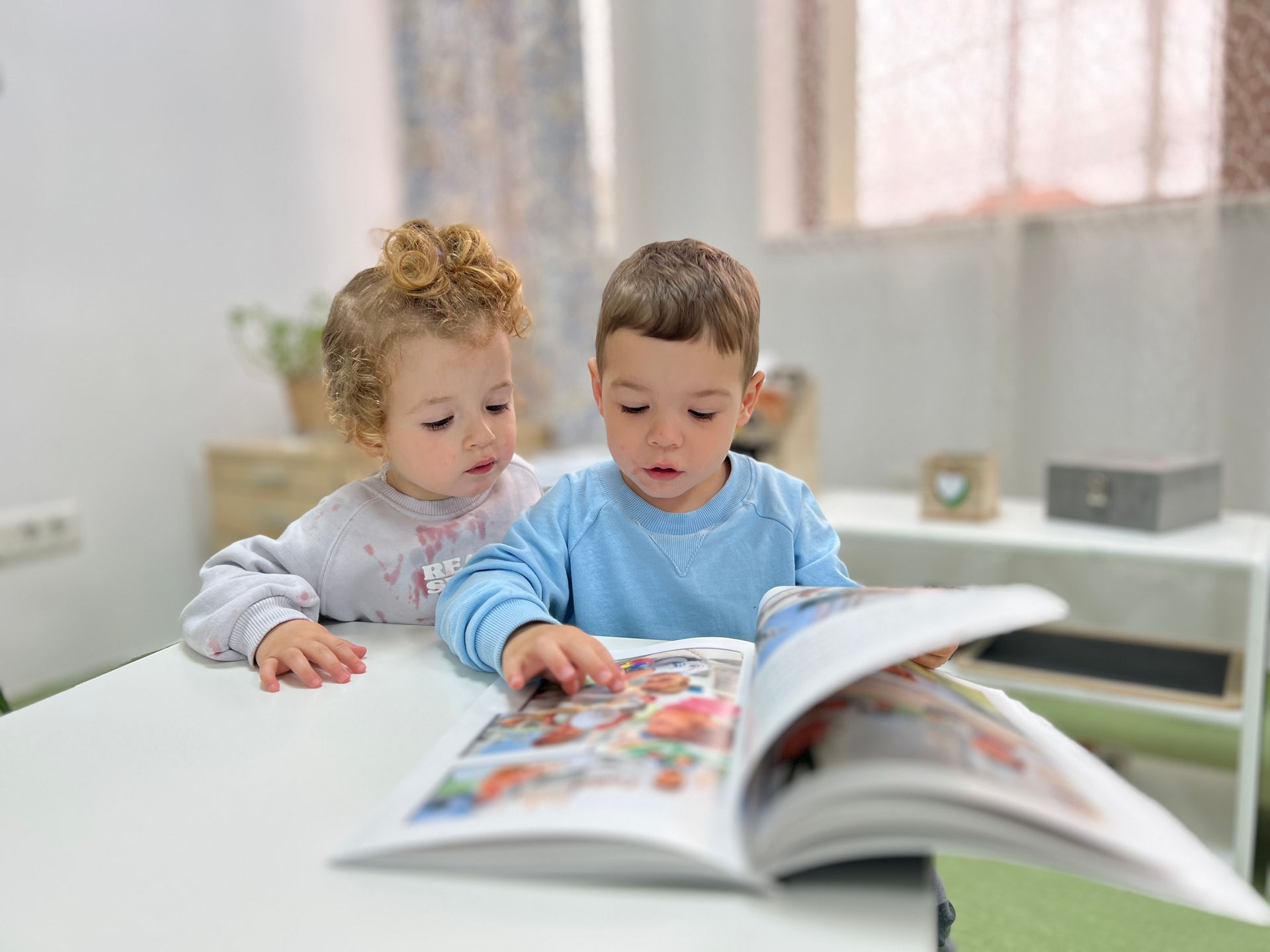 A boy and a girl are sitting at a table reading a book.