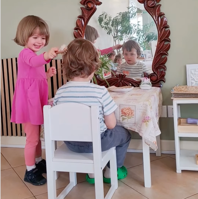 A girl in a pink dress brushes a child 's hair in front of a mirror