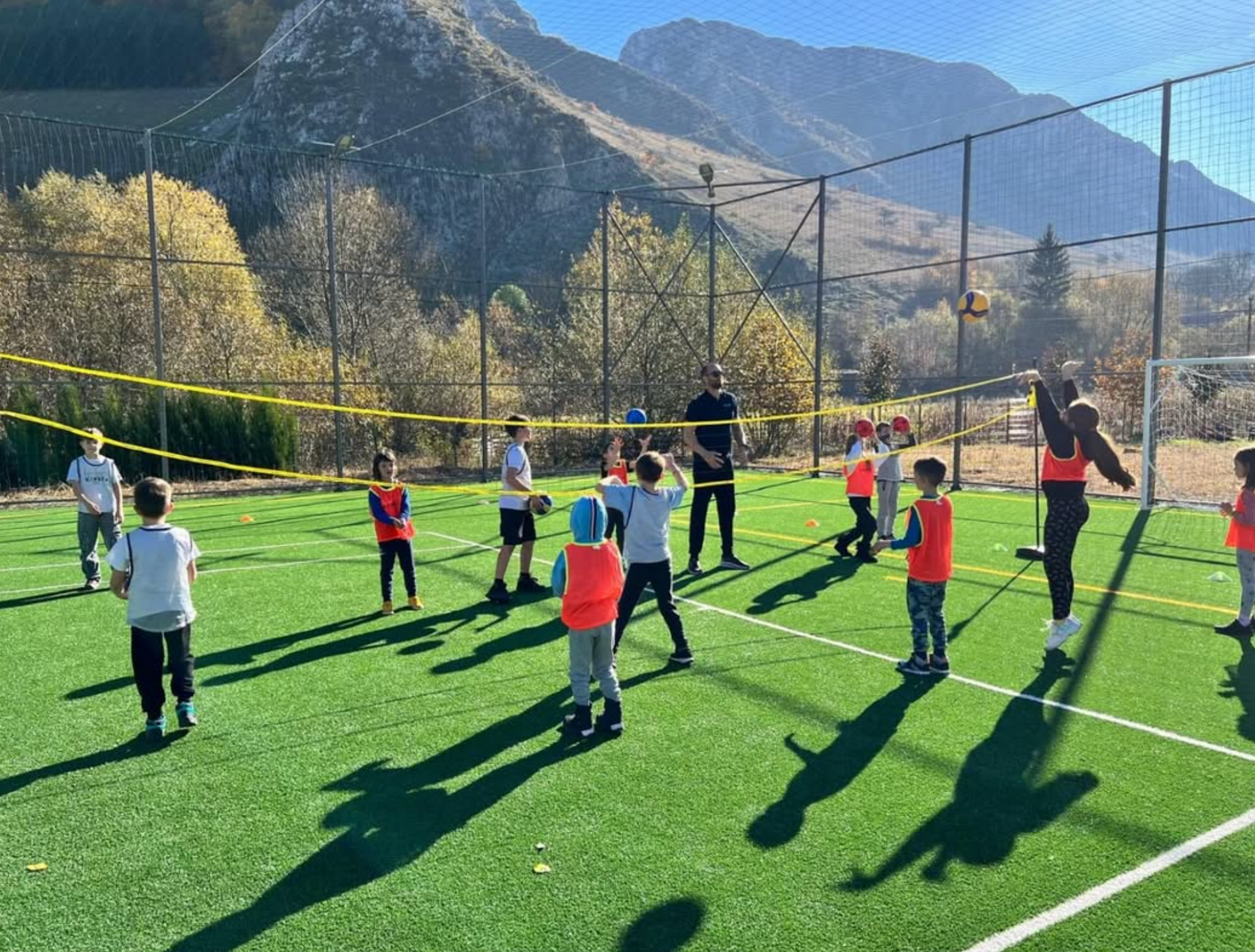 A group of children are playing volleyball on a field with mountains in the background.