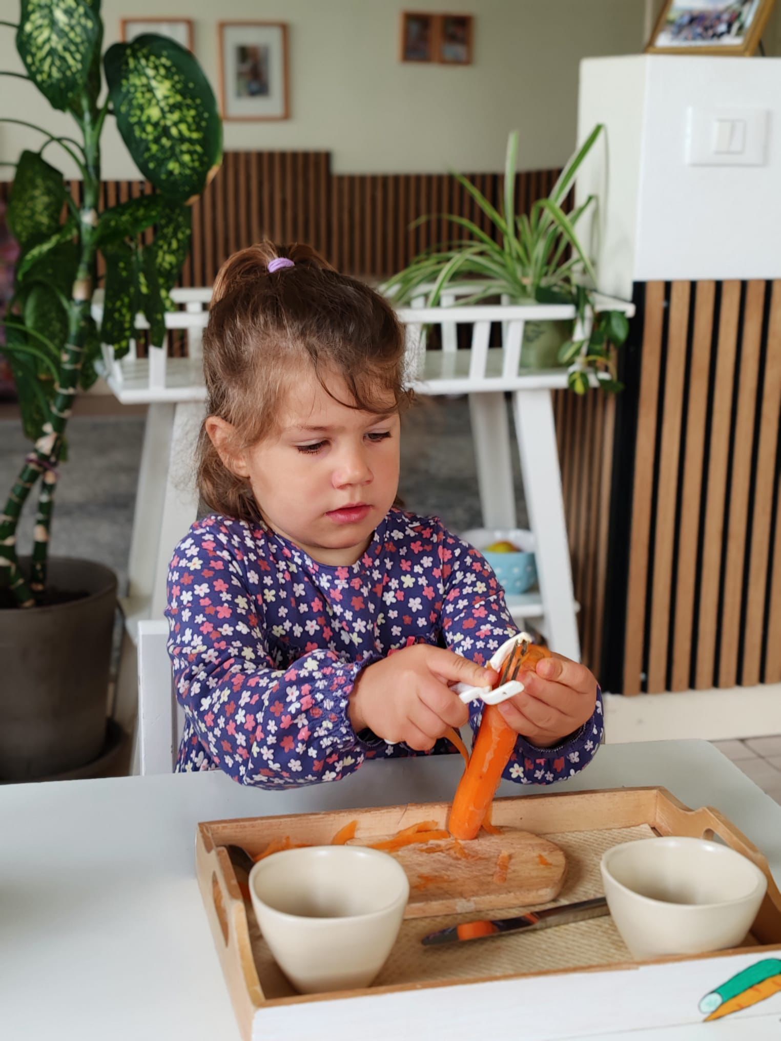 A little girl is peeling a carrot on a wooden tray.