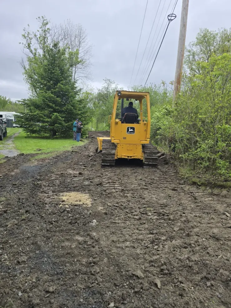 Yellow bulldozer smoothing out a muddy road, two people watching, under power lines, overcast sky.