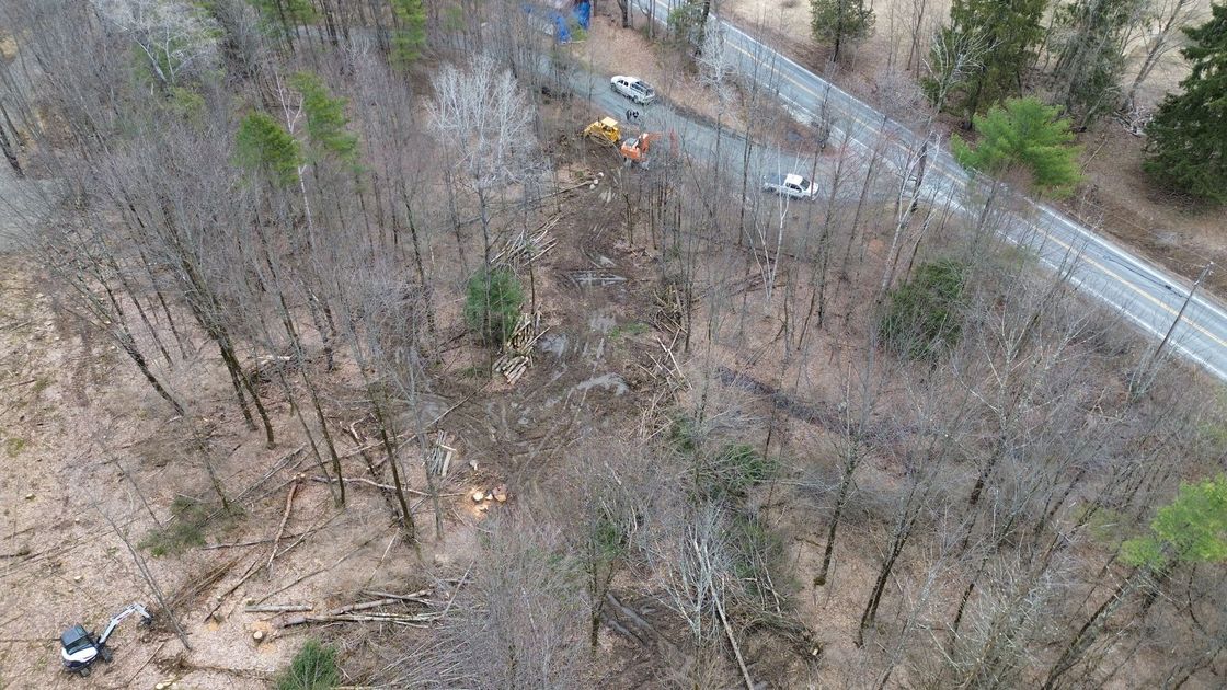 Aerial view of a wooded area beside a curving road with a small yellow excavator near the top center.