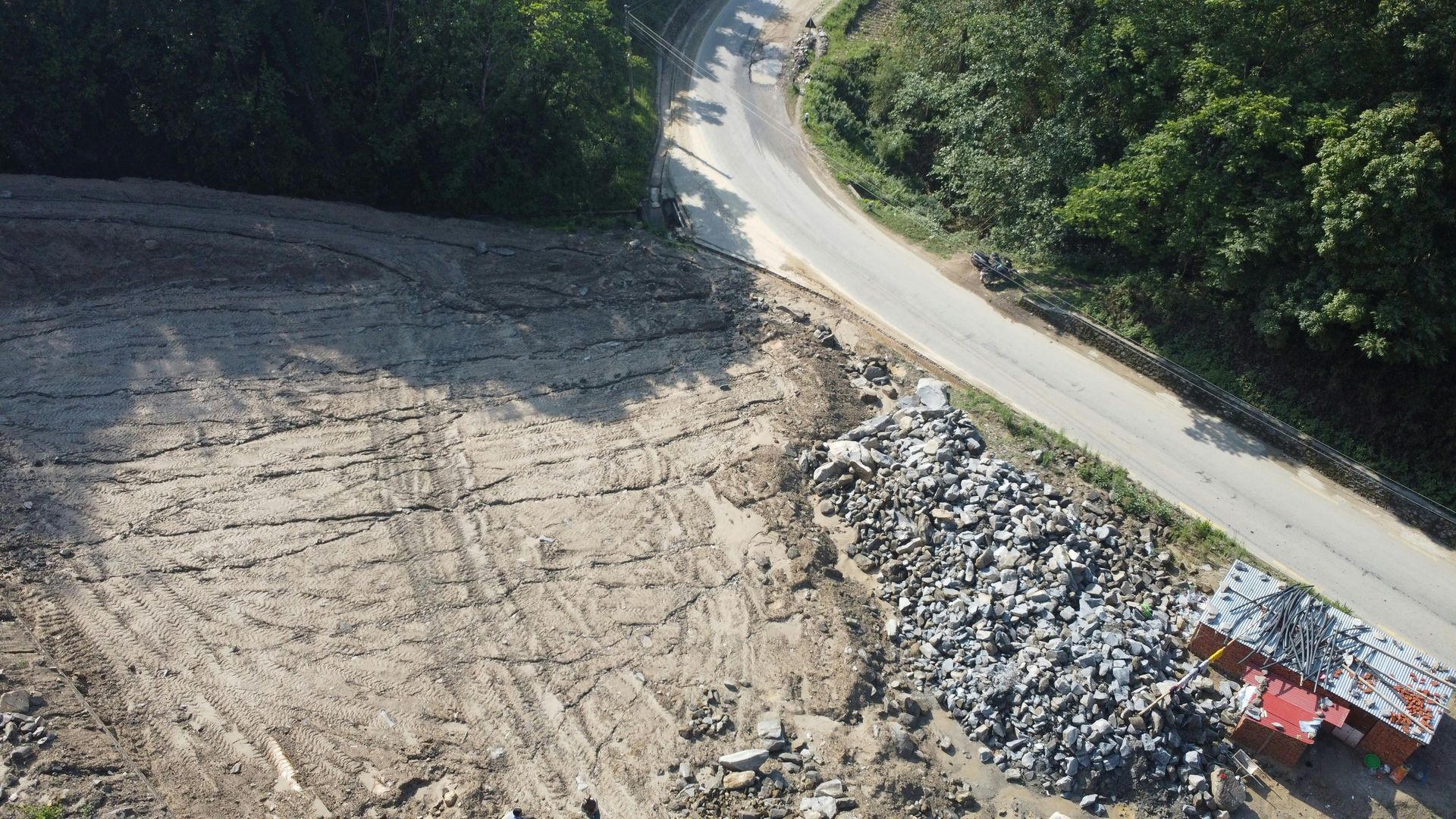 Aerial view of a dirt clearing beside a road, featuring a large pile of debris next to a small, damaged structure.